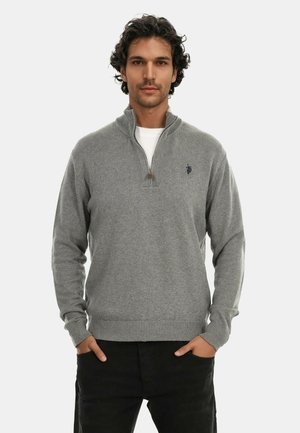 Mann mit lockigem Haar, der einen grauen Viertel-Reißverschluss-Pullover über einem weißen Hemd trägt, die Hände in den Taschen einer schwarzen Hose, stehend vor einem einfarbigen Hintergrund.