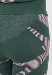 Grønne leggings med en teksturert overflate har store geometriske mønstre i lys rosa og horisontale striper på midjen.