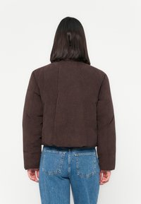 Persona con capelli lisci di lunghezza spalla di colore scuro, che indossa una giacca in velluto a coste marrone scuro e jeans di denim blu, di spalle rispetto alla macchina fotografica.