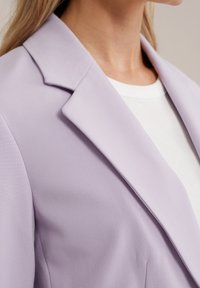 Kvinna som bär en pastell-lila blazer över en vit ribbad skjorta, som visar kragen och övre bröstområdet, neutral bakgrund.