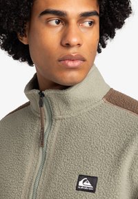 Fleece jacket in light green with a high collar, brown textured shoulder panels, zippered front, and logo patch on the chest.