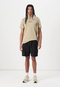 Beige bavlněné tričko s logem Timberland, černé cargo šortky, béžové tenisky s bílými akcenty, stojící na obyčejném bílém pozadí.