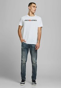 Weißes Baumwoll-T-Shirt mit einem schwarzen und roten "JACK & JONES"-Logo. Kombiniert mit ausgewaschenen blauen Jeans und schwarzen Sneakers.