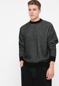 Maglione lavorato a maglia bianco e nero con un motivo testurizzato, colletto rotondo e polsini a costine, abbinato a pantaloni scuri.