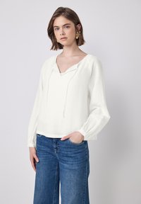 Hvid, langærmet bluse med V-udskæring og snor; har elastiske manchetter og en afslappet pasform. Bæret med blå vide jeans.
