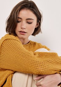 Senfgelber, gestrickter Pullover mit einem strukturierten, offenen Gewebedesign. Das Modell hat die Arme um die Knie gelegt und zeigt die lockere Passform.