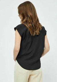 Schwarze Bluse mit kurzen Ärmeln und runder Saumform, aus weichem Material, entworfen für einen lässigen Schnitt. Getragen mit hellen Hosen.