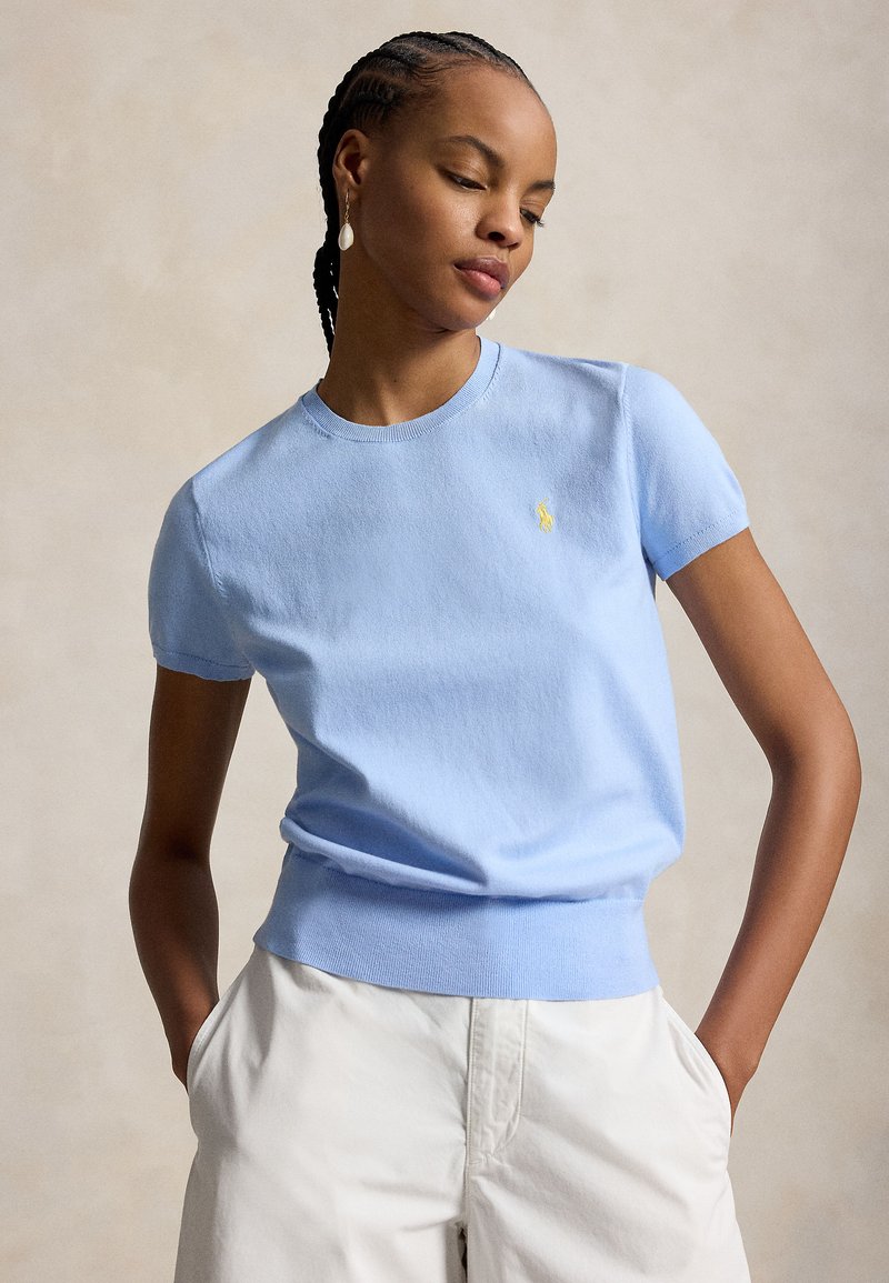 Kvinne med flettehår iført lyseblå kortermet genser og hvite bukser, med hendene i lommene, ser ned med et nøytralt uttrykk.