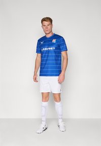 Maglia da football a maniche corte di colore blu con strisce orizzontali, loghi degli sponsor bianchi e un motivo geometrico sulle spalle, abbinata a pantaloni bianchi.