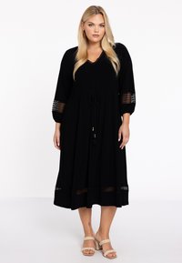 Schwarzes Maxikleid mit V-Ausschnitt, 3/4-Ärmeln und Spitzenakzenten an den Bündchen und dem Saum. Mit einem Kordelzug in der Taille und Quasten-Details.