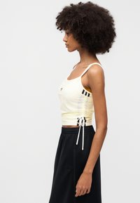 Κίτρινο crop top με ρυθμιζόμενη ρυτιδωτή λεπτομέρεια στο πλάι, λεπτές τιράντες και μαύρες διακοσμητικές ρίγες· συνδυασμένο με παντελόνια με μαύρη ελαστική ζώνη στη μέση.