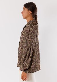 Blusa floreale marrone a vestibilità ampia, con maniche lunghe a sbuffo e chiusura con bottoni. Il tessuto ha un aspetto morbido e texturizzato.
