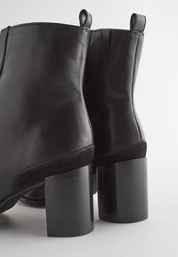 Bottines noires avec un extérieur en cuir lisse, des accents légèrement texturés et un talon bloc cylindrique robuste. Comprend des tirettes pour un enfilage facile.