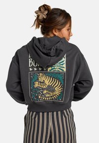 Sudadera gris oscuro con un gráfico de un tigre en la parte posterior en dorado, verde azulado y verde, con un bolsillo tipo canguro y dobladillo acanalado. Pantalones a rayas abajo.