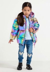 Doudoune irisée avec capuche, fermeture éclair devant, portée sur une chemise bleu clair et un jean bleu avec des patchs en forme de cœur. Bottes noires.