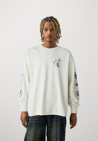Tricou cu mânecă lungă crema din bumbac, cu imprimeuri negre pe piept și mâneci. Dispune de manșete ribbate și o croială lejeră.