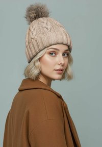 Knit beige beanie with textured cable design and a brown fur pompom on top. Worn with a brown collar coat, against a light green backdrop.