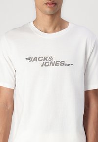 Camiseta de algodón blanca con un estampado gráfico central en gris que dice "JACK & JONES". Mangas cortas y escote redondo. Textura suave.
