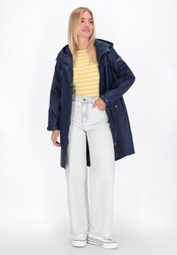 Chaqueta impermeable azul marino con capucha, botones a presión y bolsillos, combinada con una camiseta de rayas amarillas y pantalones de mezclilla claros de tiro alto.