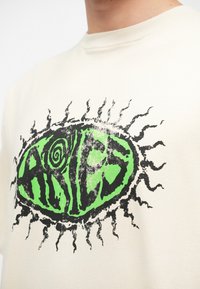 Tricou de culoare crem, cu un grafic proeminent al unui design verde, învârtejit, reprezentând "Berbec", încercuit de raze de soare negre.