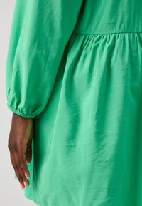 Grünes Stoffkleid mit geraffter Taille, langen Puffärmeln und glatter Textur. Der Ärmel hat einen elastischen Bund für eine gute Passform.