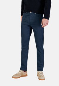 Chino blu navy slim fit con una trama liscia, cinque tasche e chiusura con bottone in metallo, abbinati a sneaker beige chiaro.