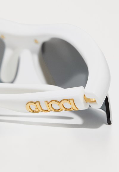 Occhiali da sole bianchi con montatura spessa, caratterizzati da un logo "Gucci" dorato sul lato e bracci neri. Texture liscia con dettagli raffinati.