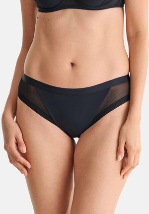 Femme portant un ensemble de lingerie bikini noir à panneaux en mesh et un soutien-gorge assorti, debout devant un fond uni.