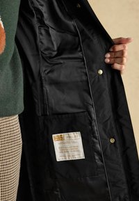 Käsi hoiab avatud musta mantlit, näidates selle siledat voodrit ning õmmeldud silti, millel on kirjutatud "Joules" ja tekst vastupidavuse ning päritolu kohta.