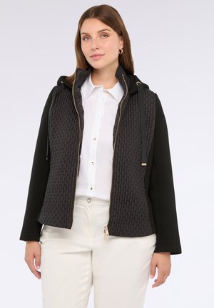 Vrouw in een zwart gewatteerd zip-up jack met capuchon over een witte blouse en lichte broek, staande tegen een witte achtergrond.
