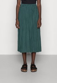 Gonna plissettata color teal con vita elasticizzata, che arriva a metà polpaccio, abbinata a sandali neri con doppia fibbia. Texture liscia.