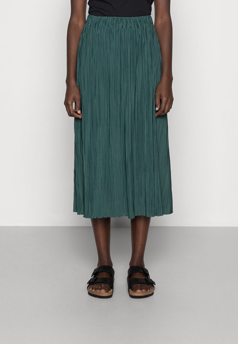 Gonna plissettata color teal con vita elasticizzata, che arriva a metà polpaccio, abbinata a sandali neri con doppia fibbia. Texture liscia.