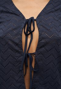 Τοπ από ναυτικό μπλε ύφασμα με οπές και ζιγκ-ζαγκ σχέδιο, δεμένο με δύο φιόγκους στο στήθος, αποκαλύπτοντας μερικά σημεία δέρματος από κάτω.