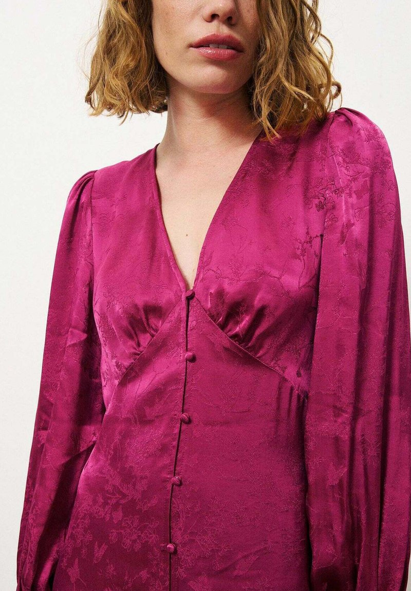 Robe en satin fuchsia avec manches longues, décolleté en v et boutons décoratifs. Présente un motif floral et une légère texture scintillante.