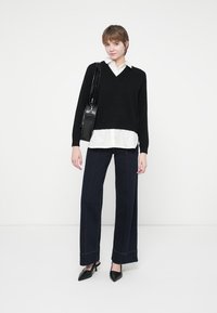 Un pull noir à col en V porté sur une chemise blanche, associé à un pantalon bleu foncé à jambes larges avec des coutures contrastantes et des escarpins noirs à bout pointu.
