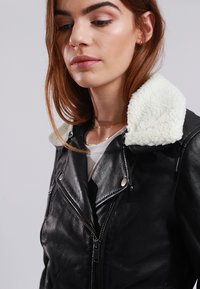 Black leather jacket with a cream faux shearling collar, zippered front, snap buttons, and a fitted design. Close-up view of the collar and lapels.