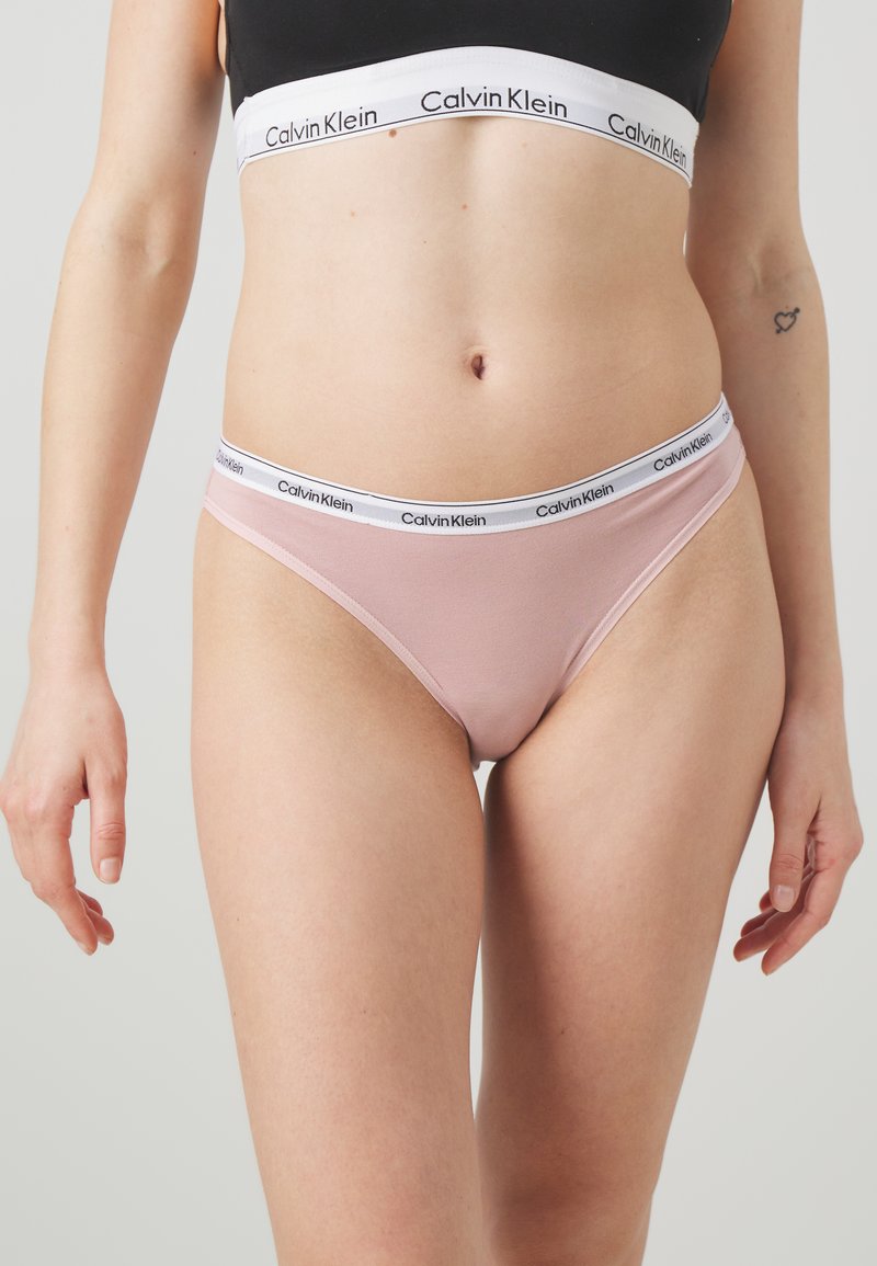 Lyserosa Calvin Klein truse med hvit elastisk midjebånd som har en svart logo. Stoffet ser glatt og elastisk ut.