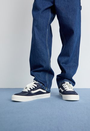 Navy sneakers with white accents, featuring textured canvas, cushioned soles, and lace-up design, paired with loose denim jeans.