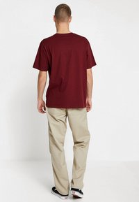 Burgunderfarbendes Baumwoll-T-Shirt mit kurzen Ärmeln, kombiniert mit beigen Hosen. Das Outfit wird mit schwarzen Sneakers abgerundet.