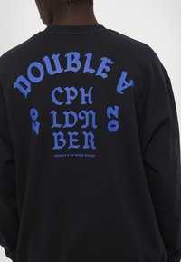 Svart genser med fet blå tekst på baksiden, med ordene "DOUBLE," "CPH," "LDN," og "BER" i en stilisert skrifttype.