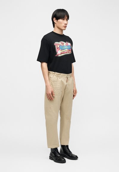Jeune homme debout de profil, portant un t-shirt noir à motif, un pantalon beige et des bottes noires à lacets sur un fond blanc uni.