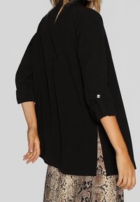 Black, long-sleeve top with rolled cuffs, featuring a loose fit and side slits. The fabric has a smooth texture. Accented with pearl buttons.
