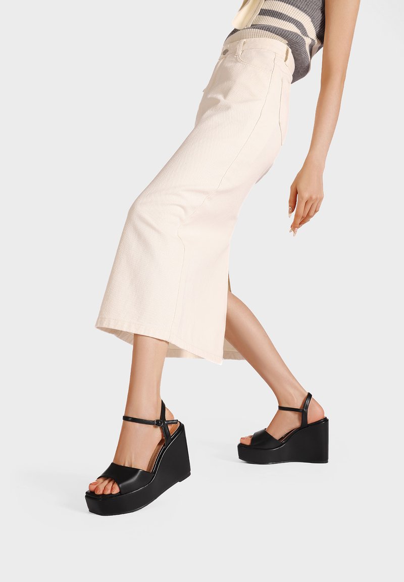 Sandali con tacco a cuneo neri con tomaia in pelle liscia, design aperto sulla punta e cinturino alla caviglia. L'altezza del tacco a cuneo è notevole. Il modello indossa una gonna pastello.