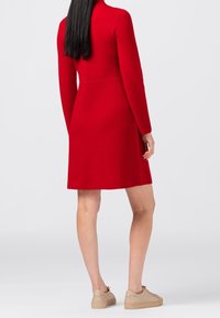 Robe rouge à manches longues avec une taille cintrée et une jupe évasée. Portée avec des baskets beiges, montrant une texture de tissu lisse.