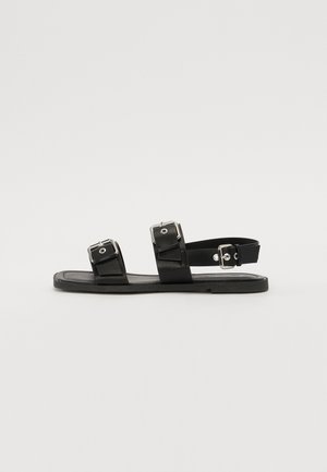 Sort flad sandal med tre justerbare remme med spænder, en over tæerne, en over mellemfoden og en rundt om anklen.