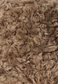 Tejido de pelo sintético rizado en marrón con un pequeño emblema decorativo metálico dorado que presenta letras entrelazadas.