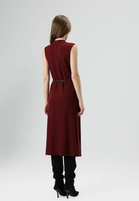 Bordeaux mouwloze jurk met een riem, zichtbare achterrits en vloeiend ontwerp, gecombineerd met zwarte nauwsluitende broek en laarzen met hoge hakken.