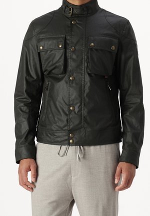 Homme portant une veste en cuir noire avec des poches à rabat boutonnées et des poches latérales zippées, par-dessus un pantalon gris clair à cordon de serrage.