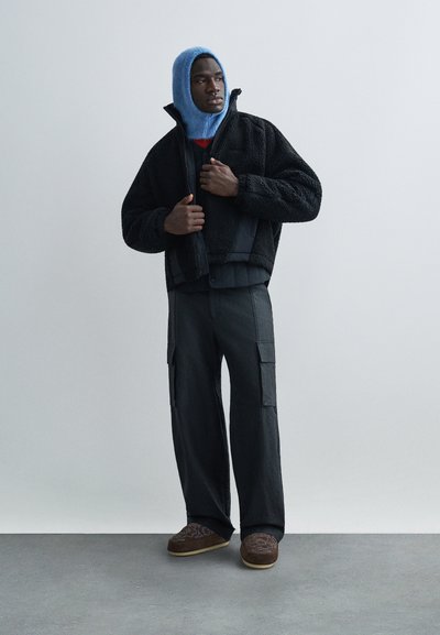 Man justerar svart fleecetröja över vadderad väst, bär blå stickad huva, lösa svarta cargobyxor och bruna slip-on skor, stående på grått golv.