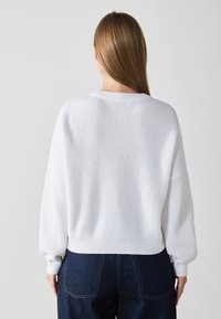 Pull en tricot blanc avec une texture côtelée et des épaules tombantes, doté un ourlet court et de larges manches, assorti à un jean en denim foncé.